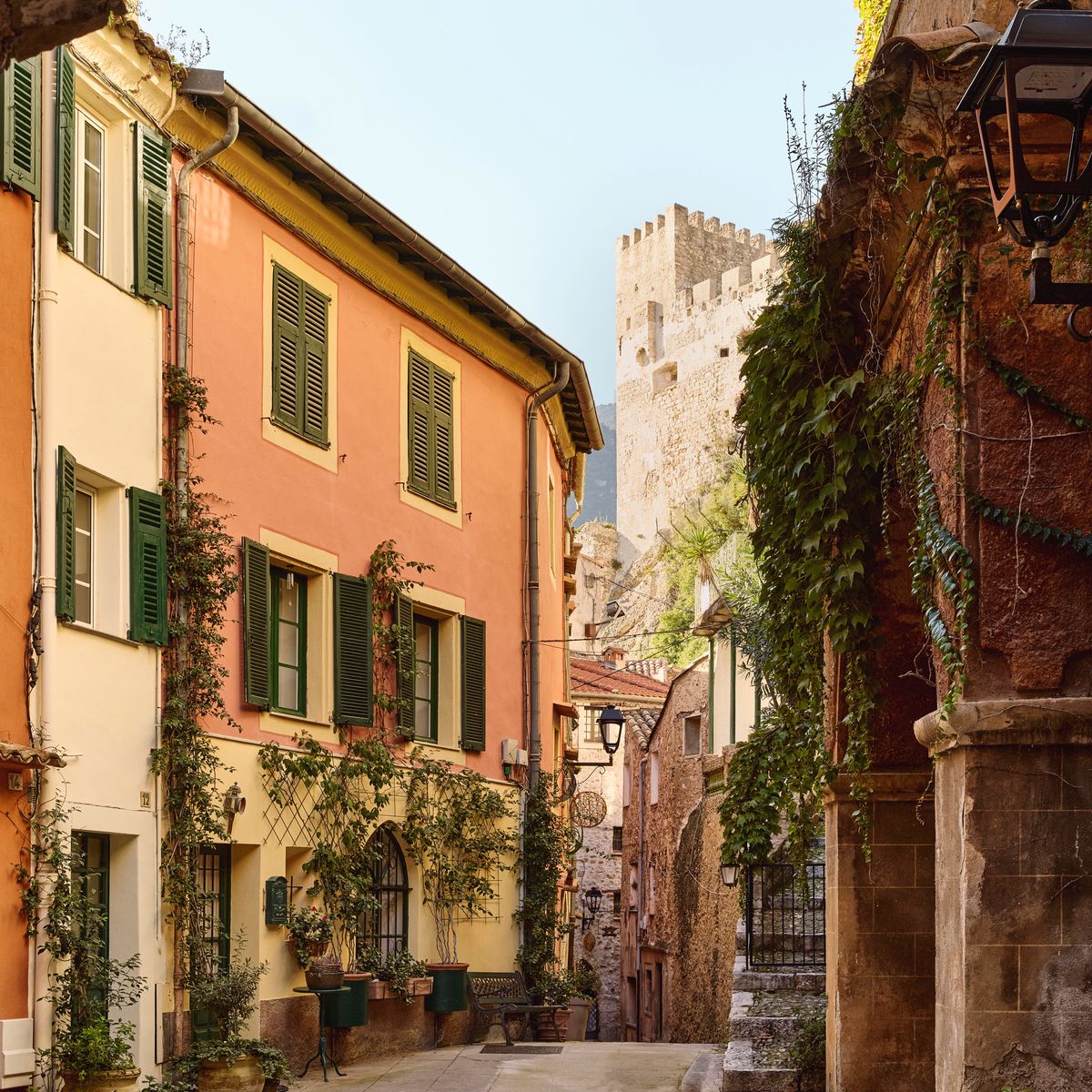 Charming narrow street with pastel buildings, green shutters, potted plants, and a distant medieval stone tower framed by an archway, under clear daylight.