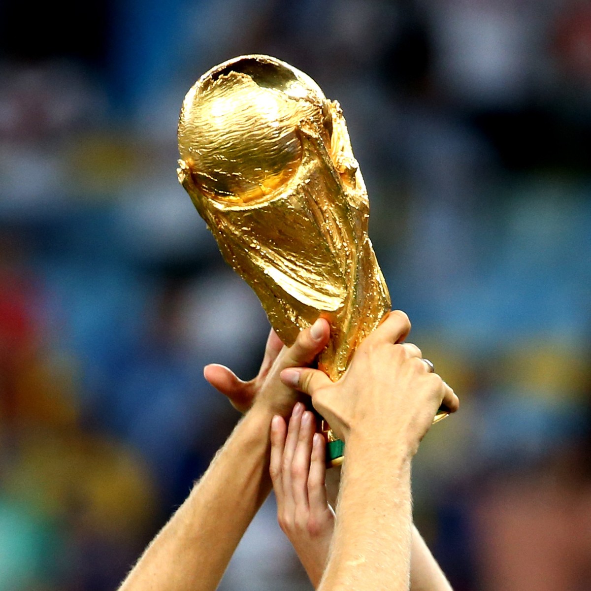 Multiple hands raise the golden FIFA World Cup trophy in celebration, with a blurred stadium background.
