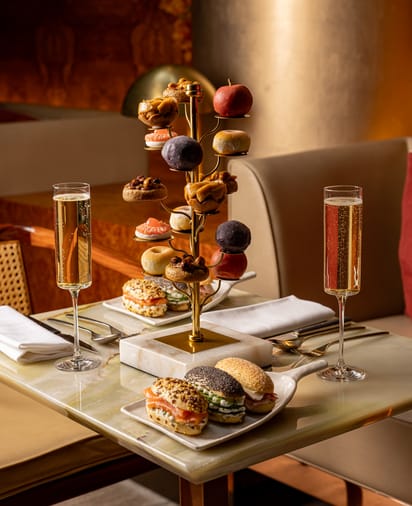 Afternoon tea with fruit-shaped pastries on a golden tree stand, gourmet sandwiches, and two glasses of sparkling wine.