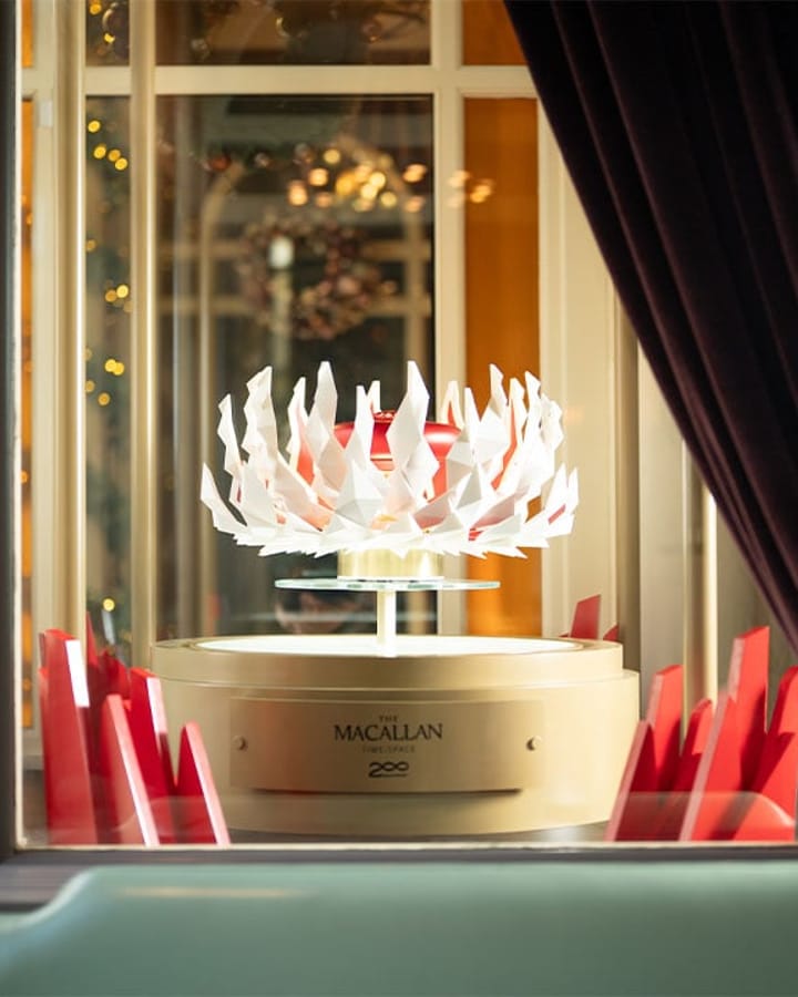 A sculptural Macallan whisky display with intricate white and red geometric elements, elegantly showcased in a glass case with ambient lighting.