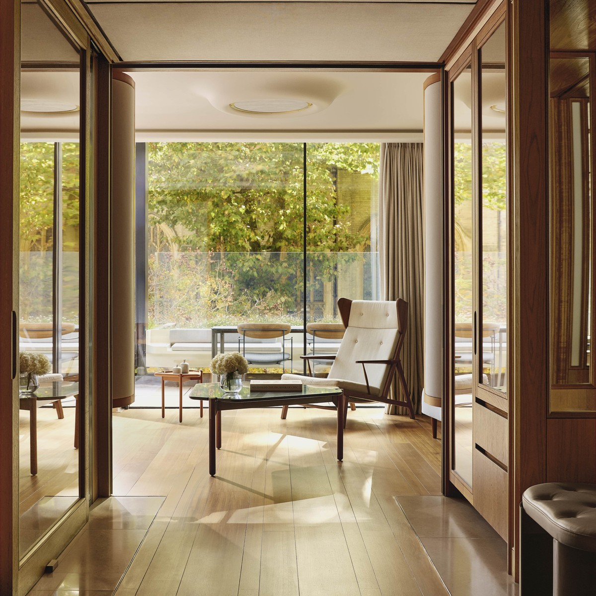 Minimalist sitting area with natural wood flooring, wood furnishings, a white cushioned loungechair, a glass top table featuring white flowers in a glass vase, and floor-to-ceiling windows opening to a private terrace in the background.