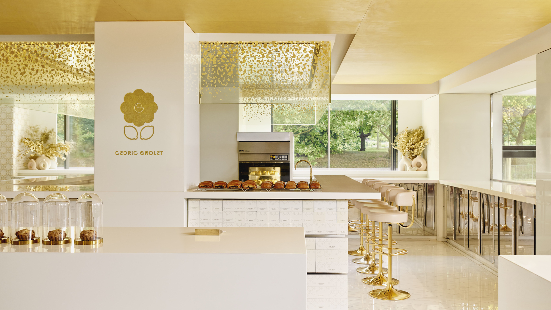 Bright, modern patisserie interior featuring a central white and gold counter with pastries under glass domes, gold-accented bar stools, and large windows overlooking greenery.