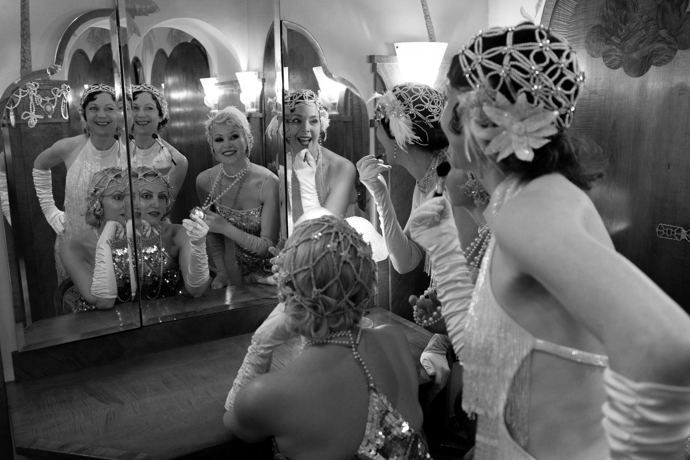 Women in 1920s costumes apply makeup and laugh together at a mirrored vanity.