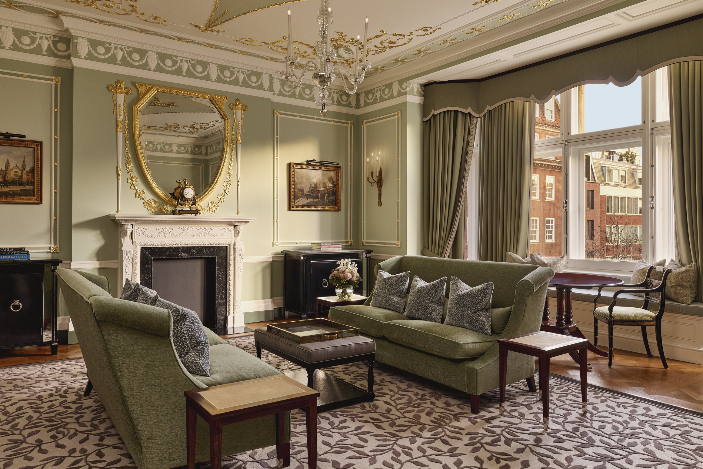 Refined suite with green sofas, ornate gold-trimmed walls, and large windows overlooking brick buildings..