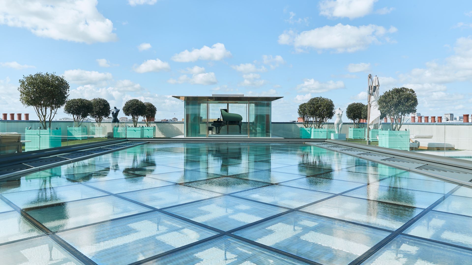 A rooftop glass pavilion with a grand piano, surrounded by reflective water features, manicured greenery, and a backdrop of blue skies with fluffy clouds.