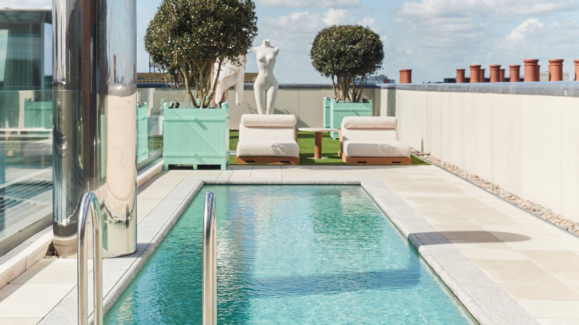 A serene rooftop pool with crystal-clear water, lined with plush loungers, sculptural art, and modern greenery under a bright blue sky.