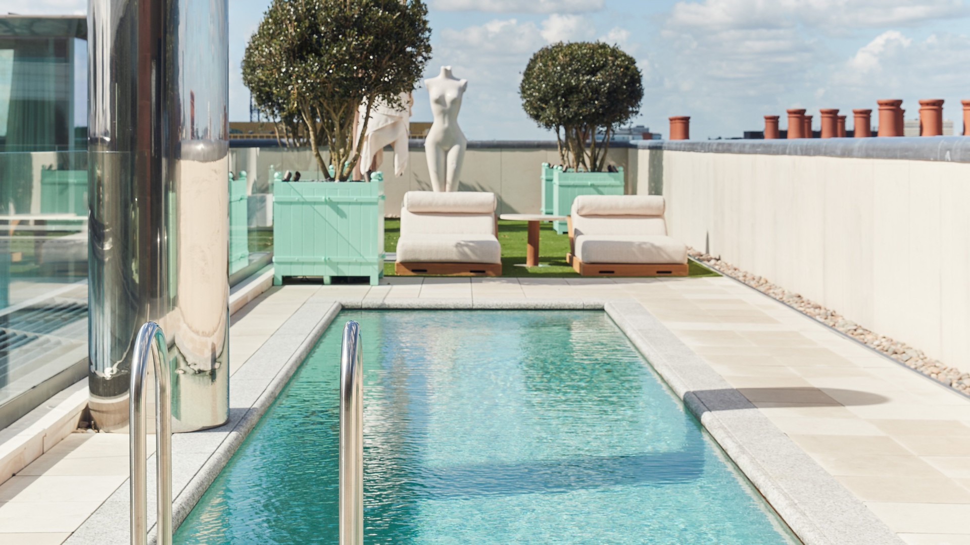 A serene rooftop pool with crystal-clear water, lined with plush loungers, sculptural art, and modern greenery under a bright blue sky.