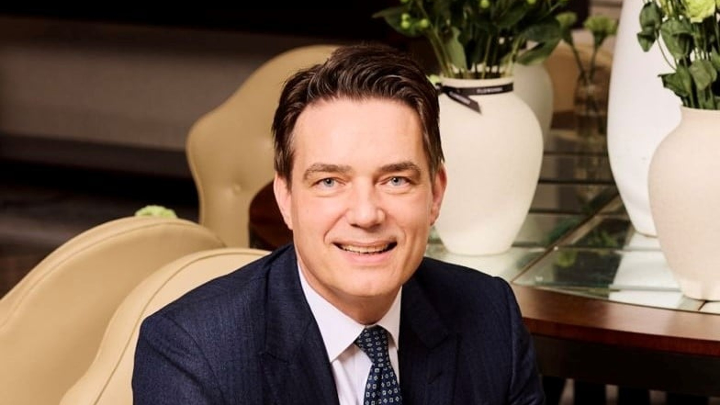 Man in a dark suit seated on a beige chair, smiling, with white vases and green flowers in the background