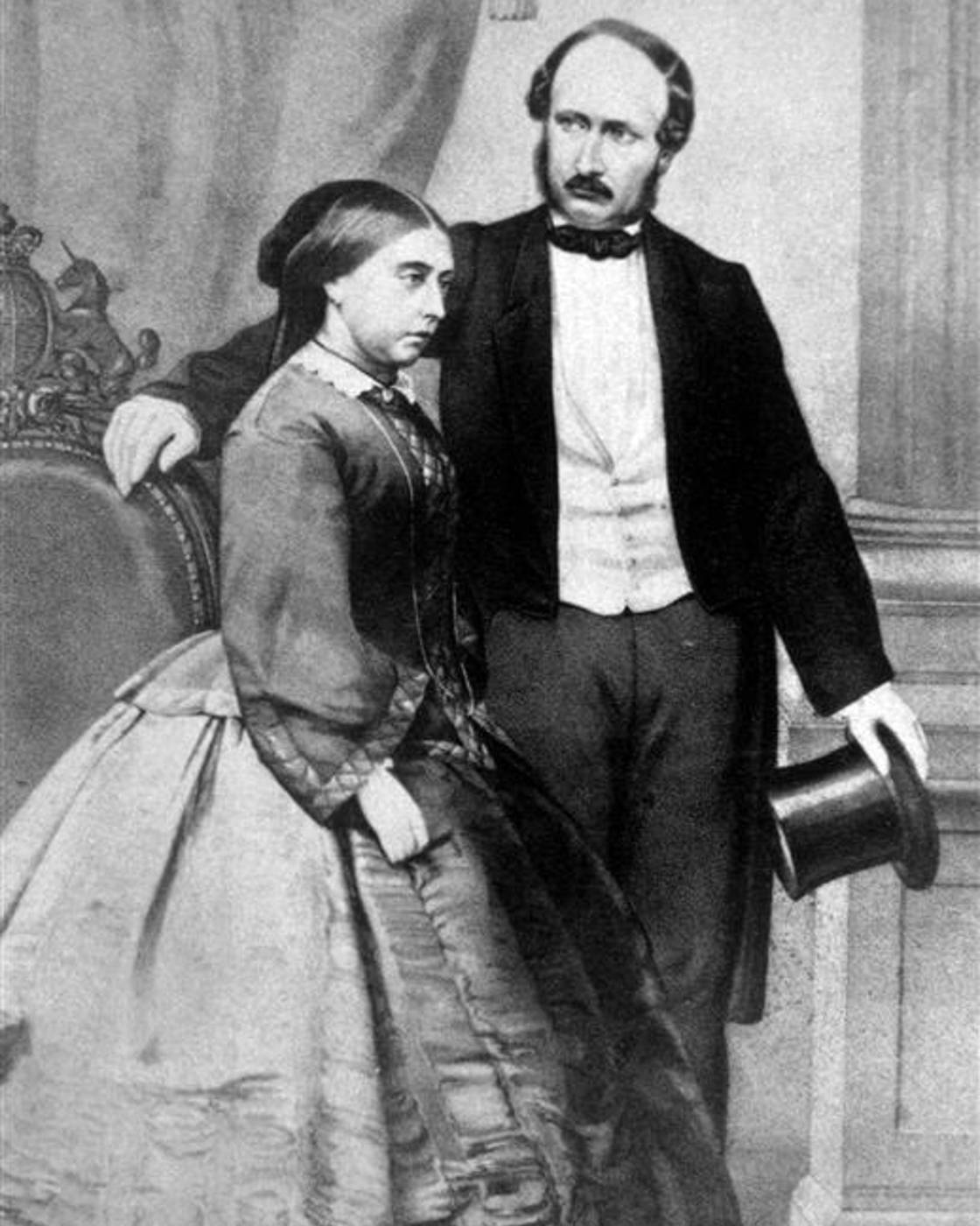 Black and white portrait of a Victorian-era couple; the woman is seated in a full gown, the man stands beside her holding a top hat.