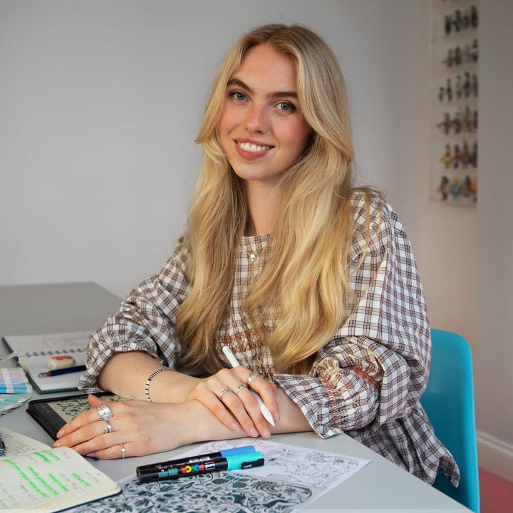 Smiling woman with long blonde hair sits at a desk with notebooks, pens, and artwork, appearing creative and focused.
