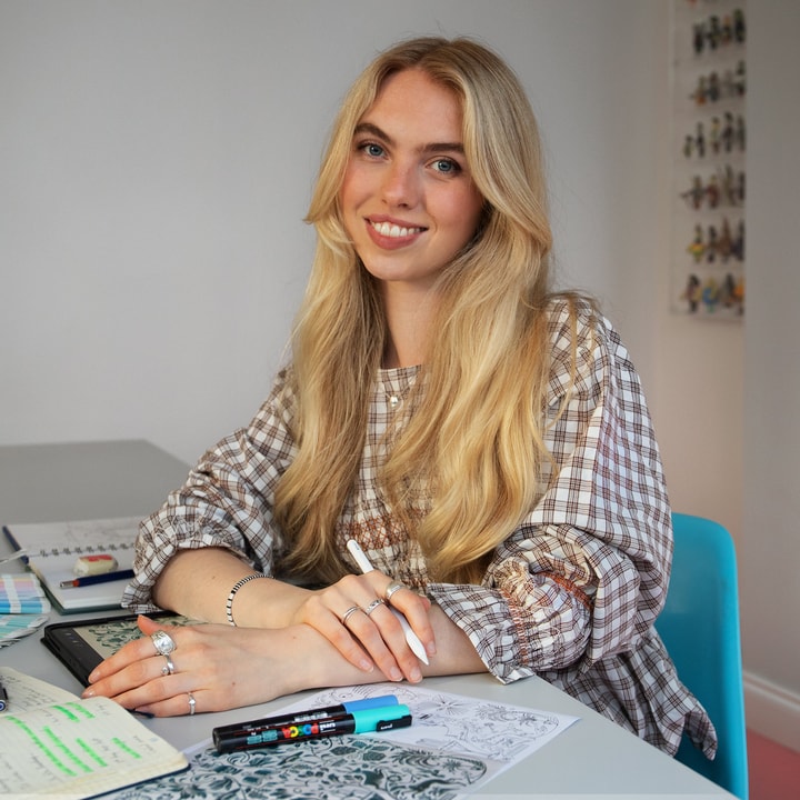 Smiling woman with long blonde hair sits at a desk with notebooks, pens, and artwork, appearing creative and focused.