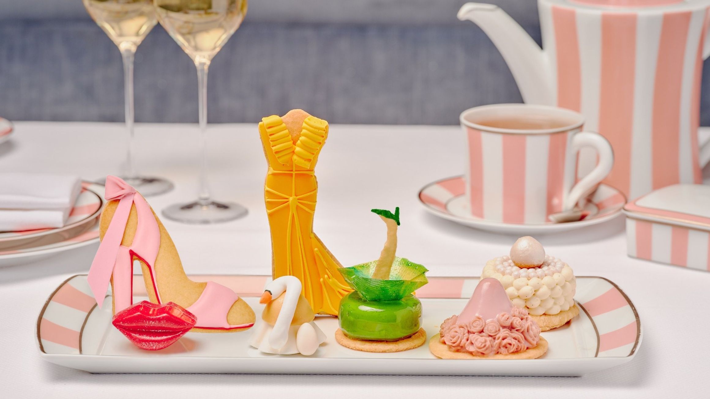 A selection of couture-inspired pastries on a rectangular plate, including a pink stiletto cookie, a yellow gown-shaped dessert, and other intricately crafted treats, served alongside striped pink-and-white teaware and two glasses of champagne.