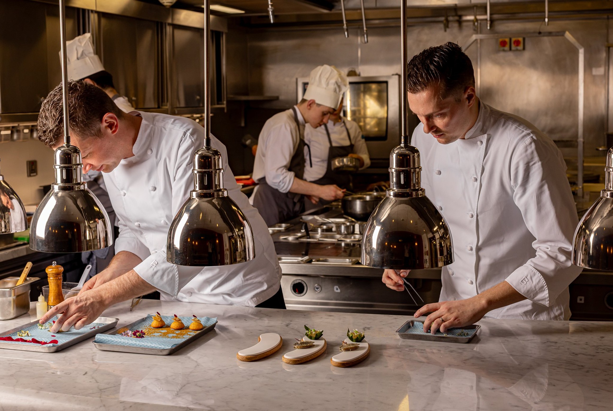 Chefs in white uniforms meticulously plating gourmet dishes under heat lamps in a professional stainless steel kitchen, with colleagues preparing food in the background.