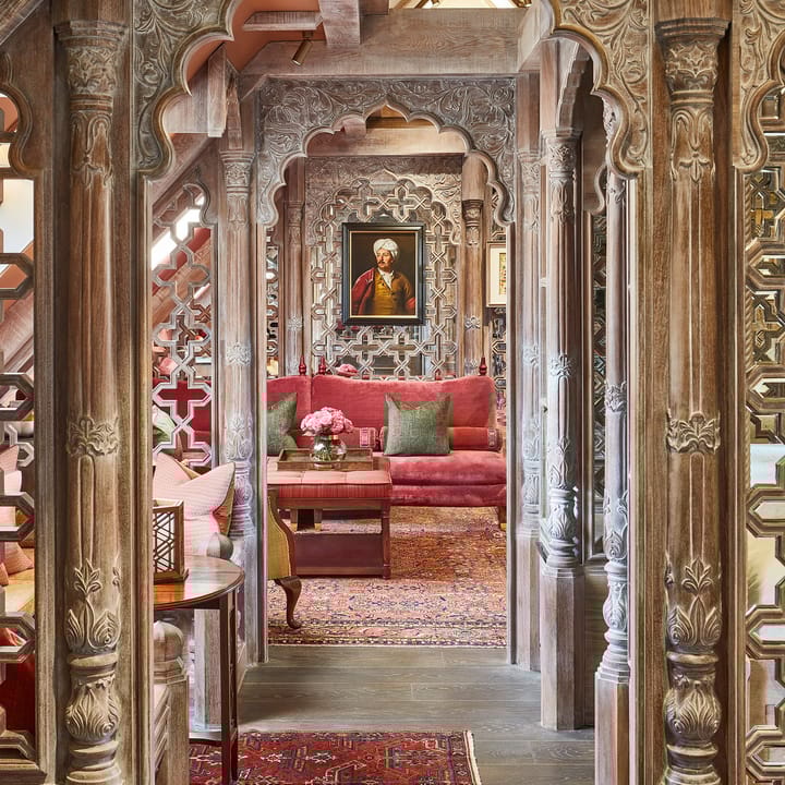 Ornate wooden corridor with carved arches leading to a richly decorated lounge with red sofa, Persian rug, and portrait.