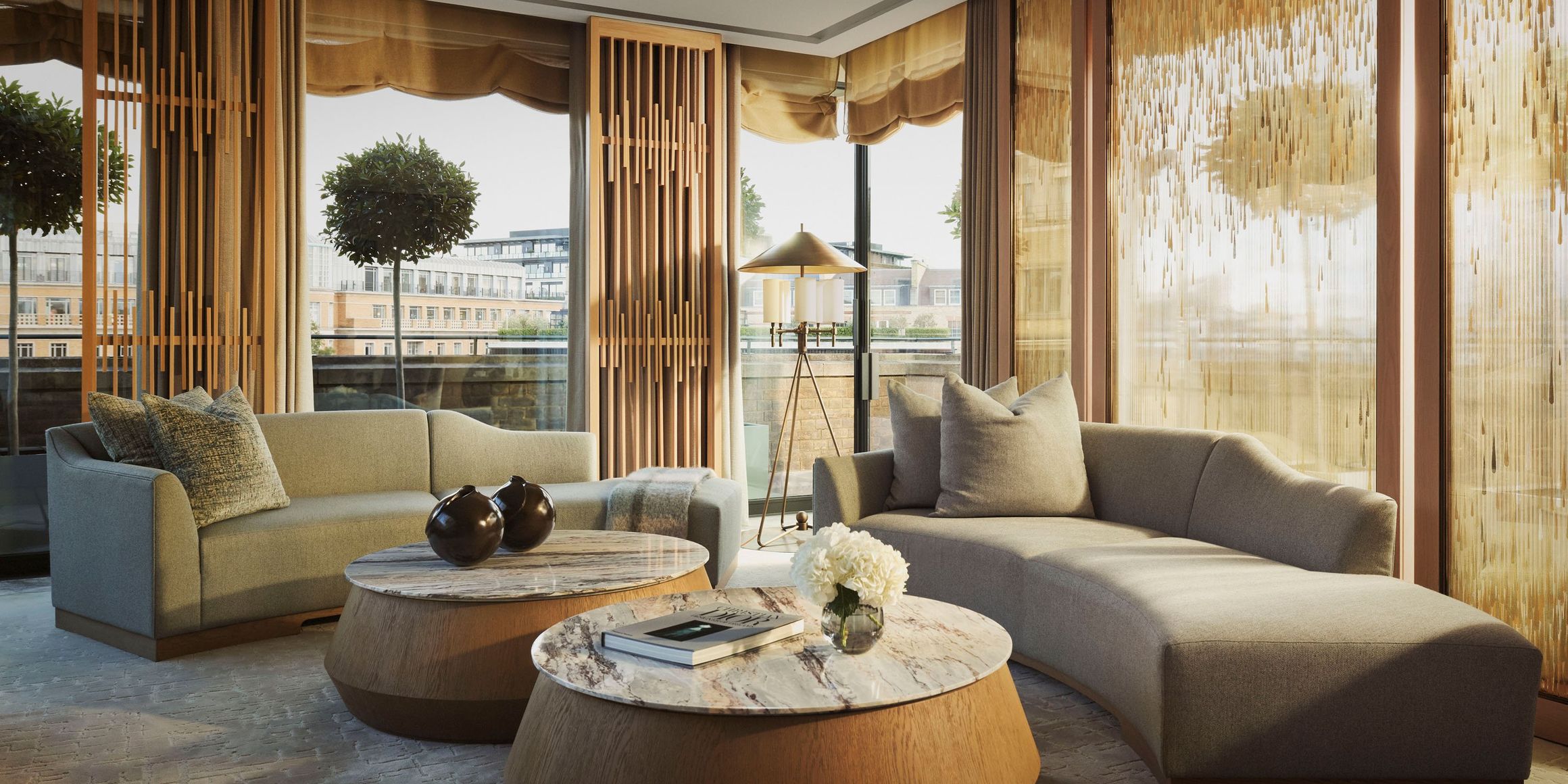 Contemporary penthouse lounge with curved sofas, marble tables, and golden-accented screens, overlooking a private terrace with topiary trees and city views.