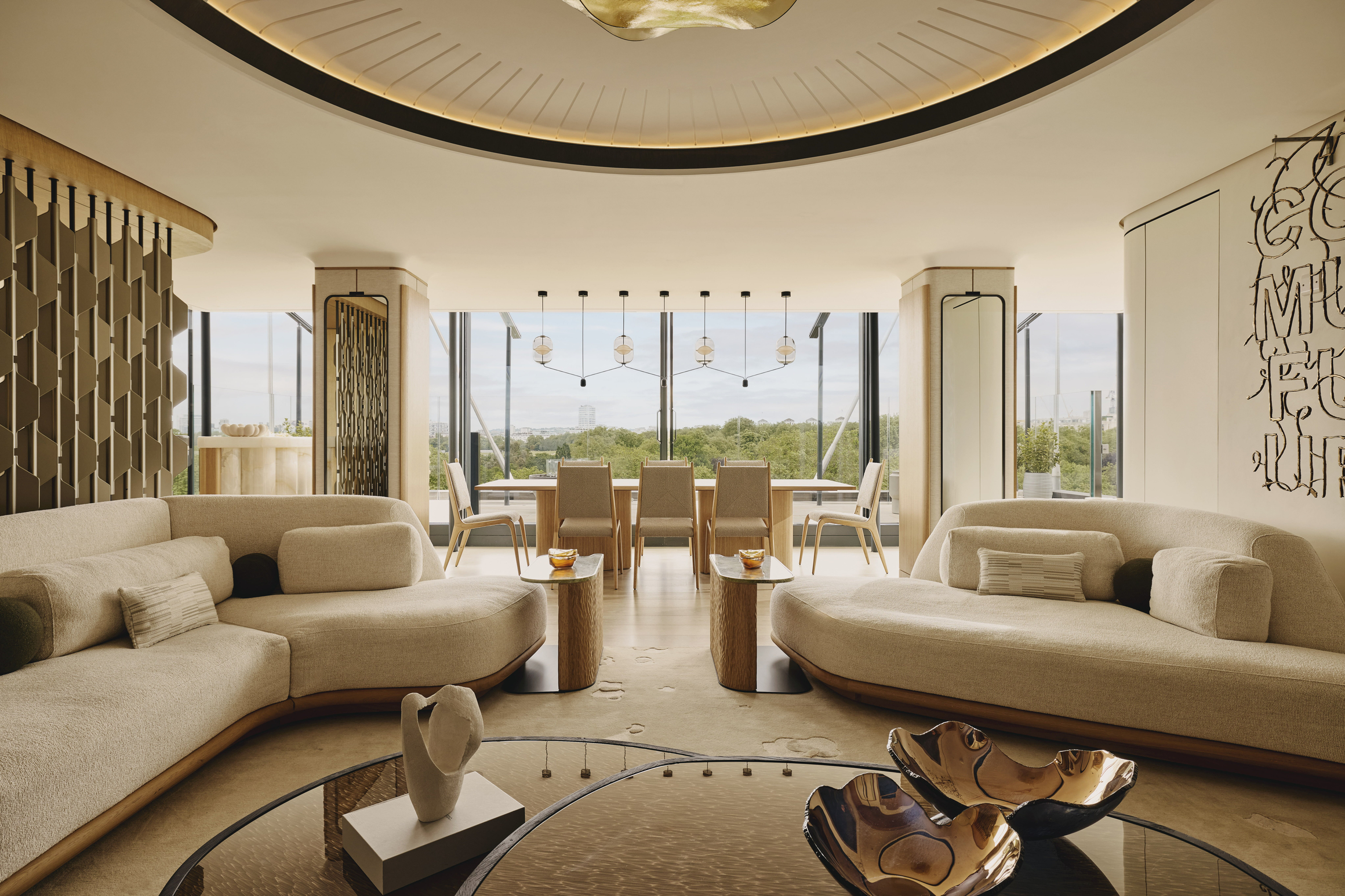 Spacious penthouse interior with sculpted cream sofas, circular ceiling detail, modern art, and full-height windows overlooking treetops.