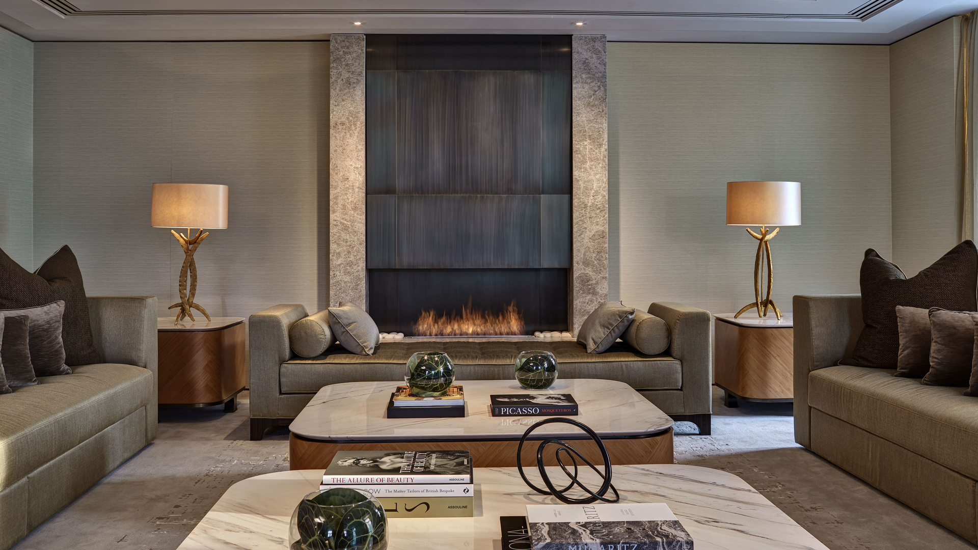 A sophisticated lounge area with plush seating, marble tables, and an elegant fireplace as the focal point. Soft lighting and tasteful decor, including art books and sculptural accents, create an inviting and luxurious atmosphere.