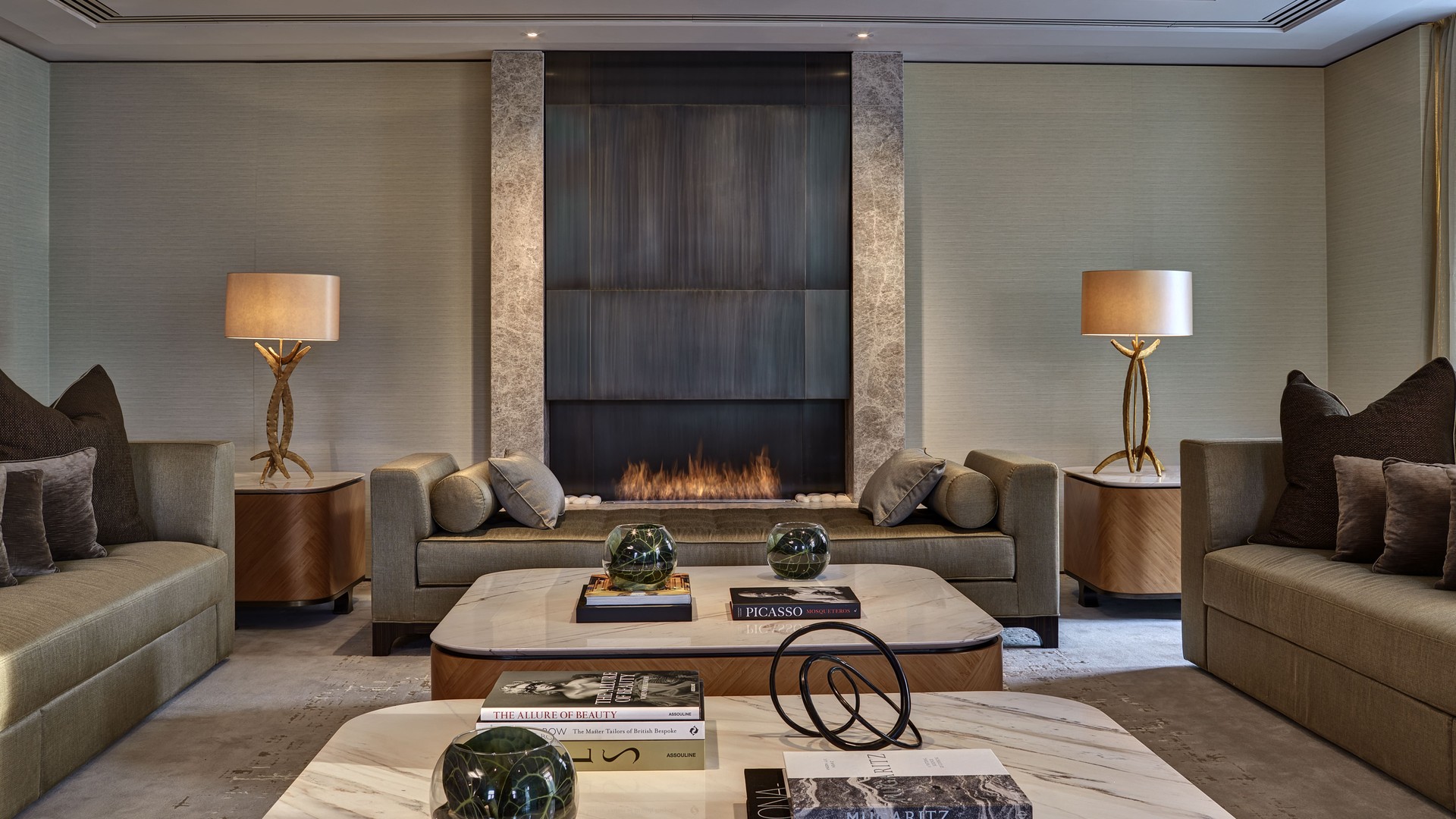 A sophisticated lounge area with plush seating, marble tables, and an elegant fireplace as the focal point. Soft lighting and tasteful decor, including art books and sculptural accents, create an inviting and luxurious atmosphere.
