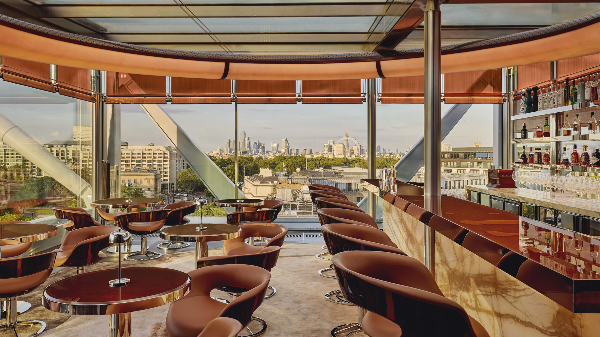 Elegant rooftop bar with panoramic city views, showcasing curved leather chairs, glossy round tables, and a marble-topped bar beneath a glass ceiling and warm-toned blinds.