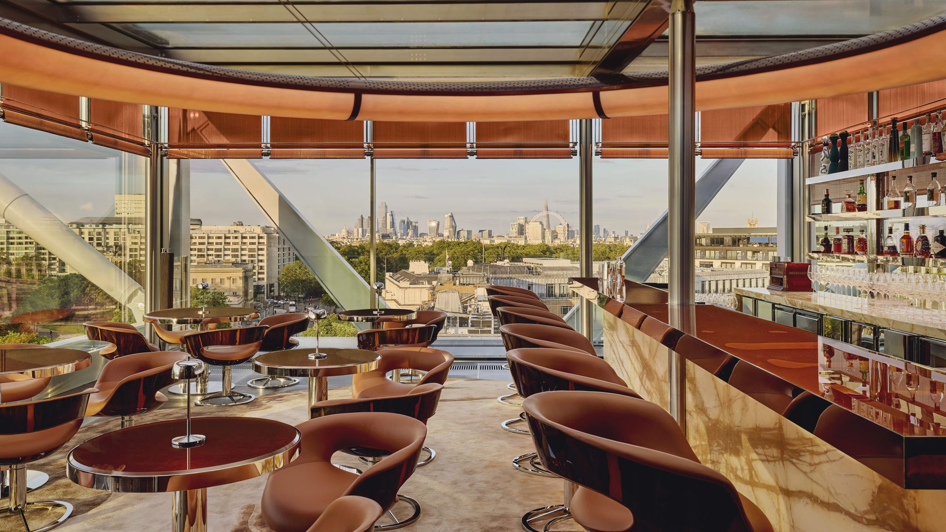 Sophisticated rooftop bar interior with sculptural seating, polished round tables, and a glowing onyx bar, framed by panoramic views of London’s skyline through floor-to-ceiling glass.