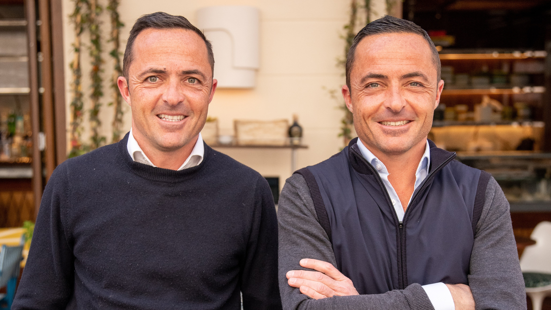 Two individuals standing side by side in a casual outdoor setting, both dressed in smart yet relaxed attire, smiling warmly towards the camera with a backdrop of greenery and a stylish café interior.