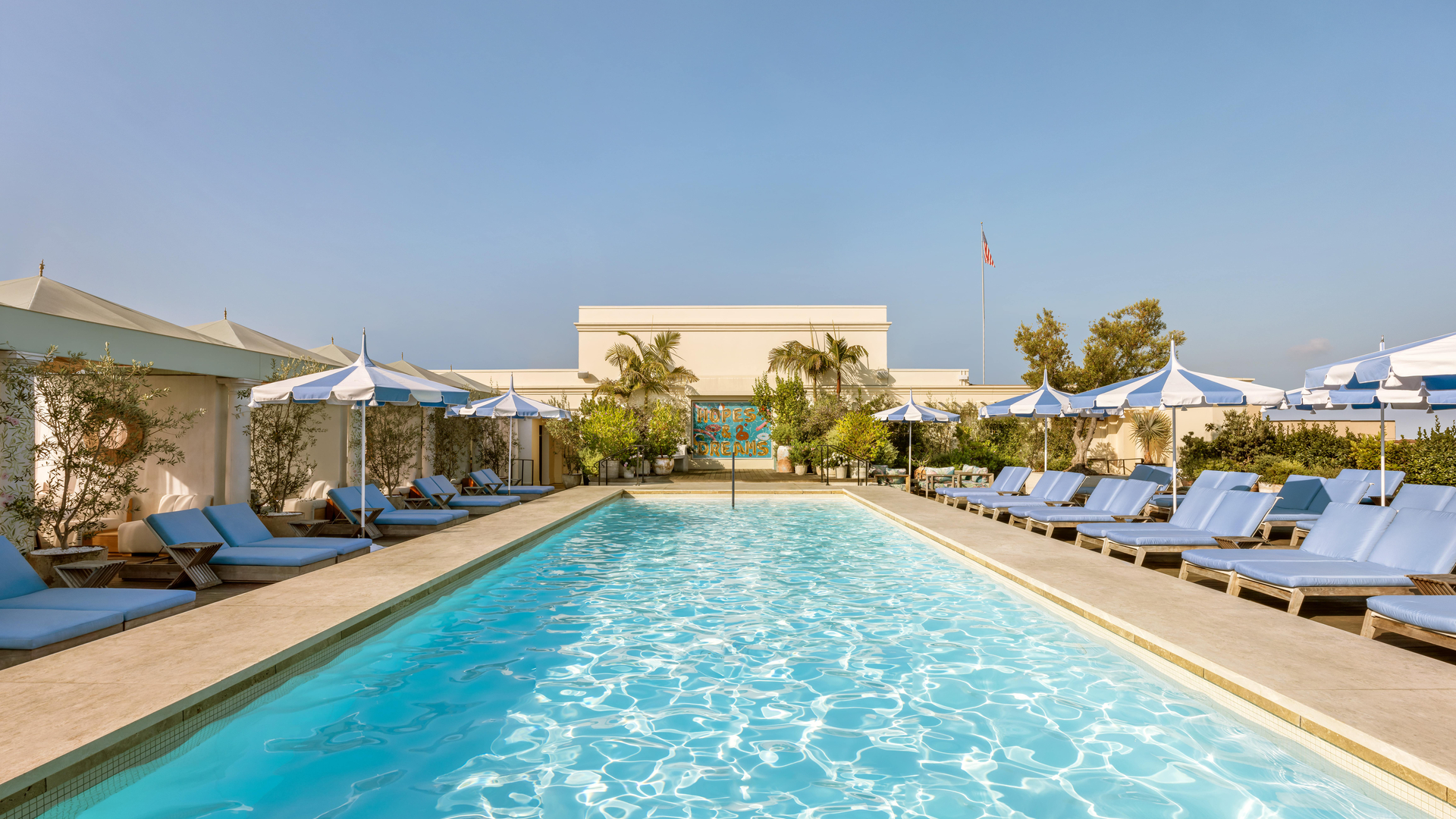 Sunlit rooftop pool lined with blue loungers, striped umbrellas, and lush greenery beneath a clear blue sky.