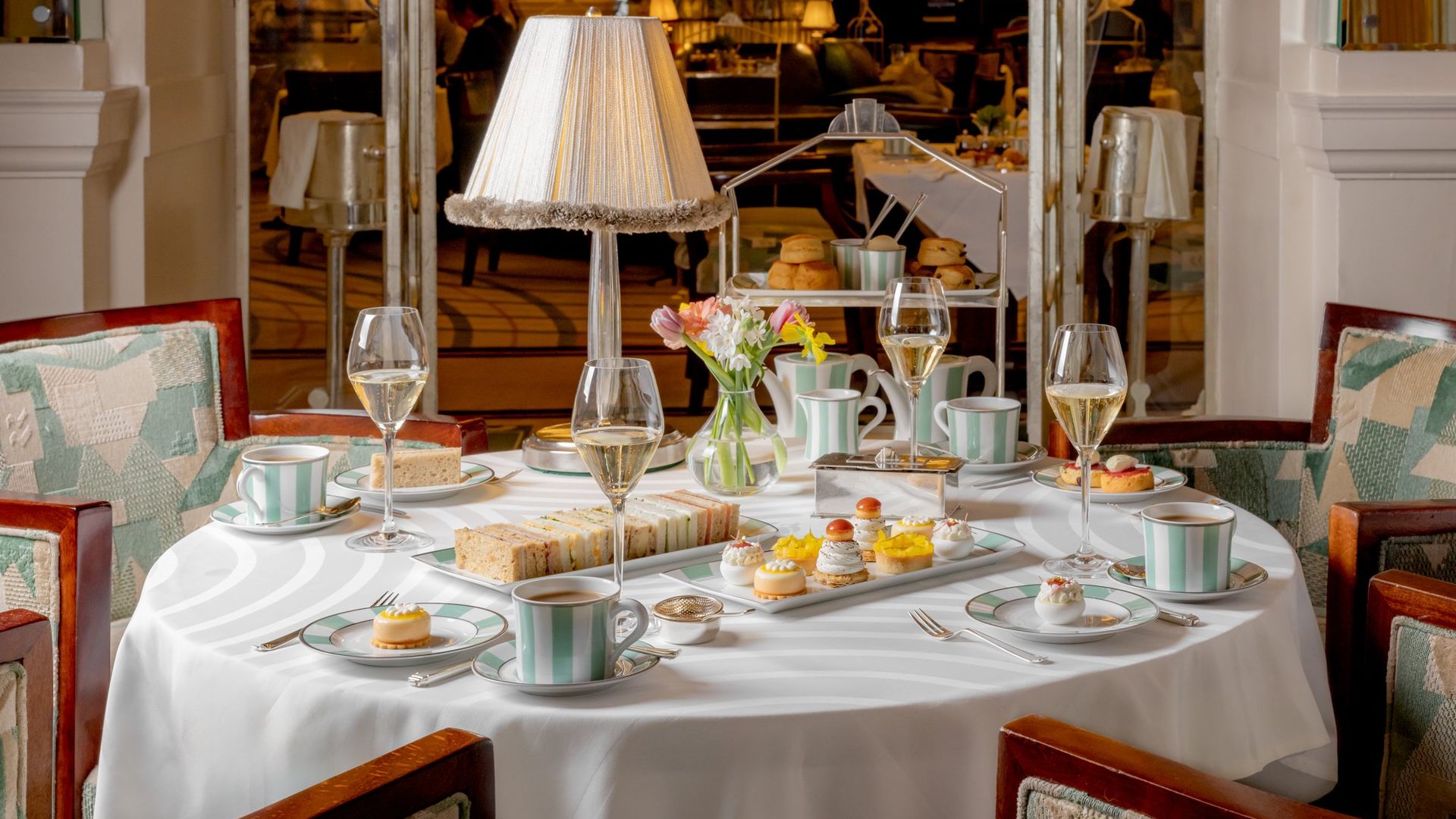 The elegant Claridge’s tea service with striped green-and-white porcelain, an assortment of finger sandwiches, delicate pastries, and sparkling wine in a grand dining space.