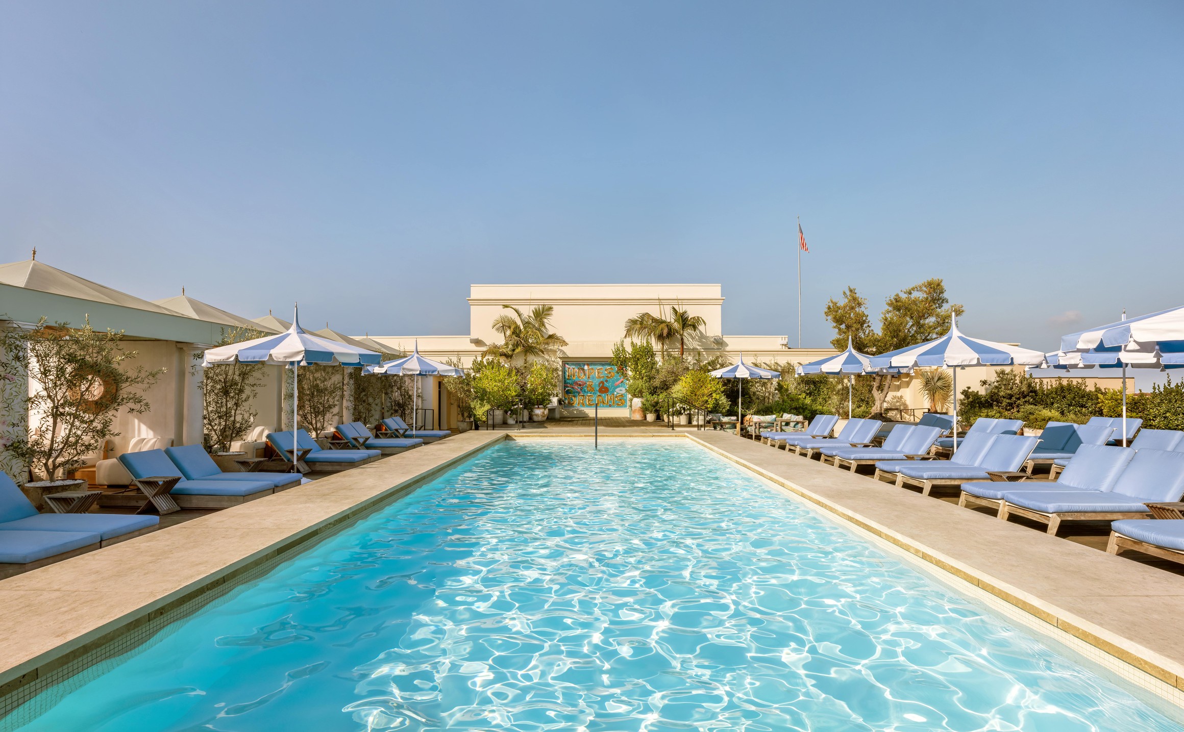 Sunlit rooftop pool lined with blue loungers, striped umbrellas, and lush greenery beneath a clear blue sky.