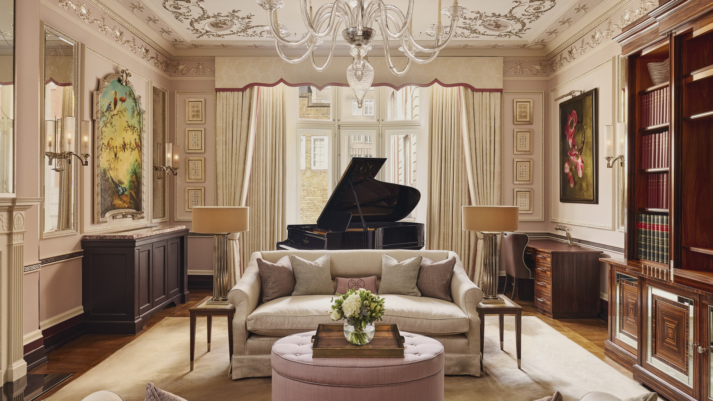 Sophisticated room with cream sofas, a grand piano, pink accents, and floral artwork under a chandelier.