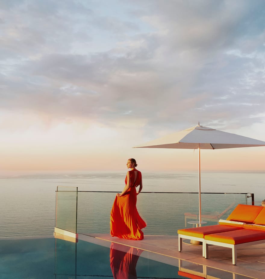 A woman in a flowing red dress gazes at the ocean from an infinity pool, bathed in golden sunset light.