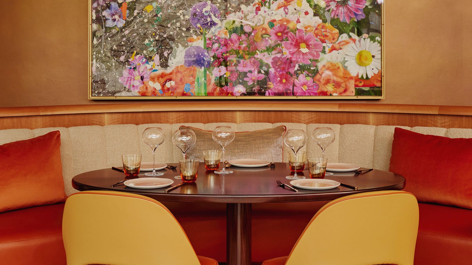 Vibrant dining nook with orange seating, glassware, and a large floral artwork.