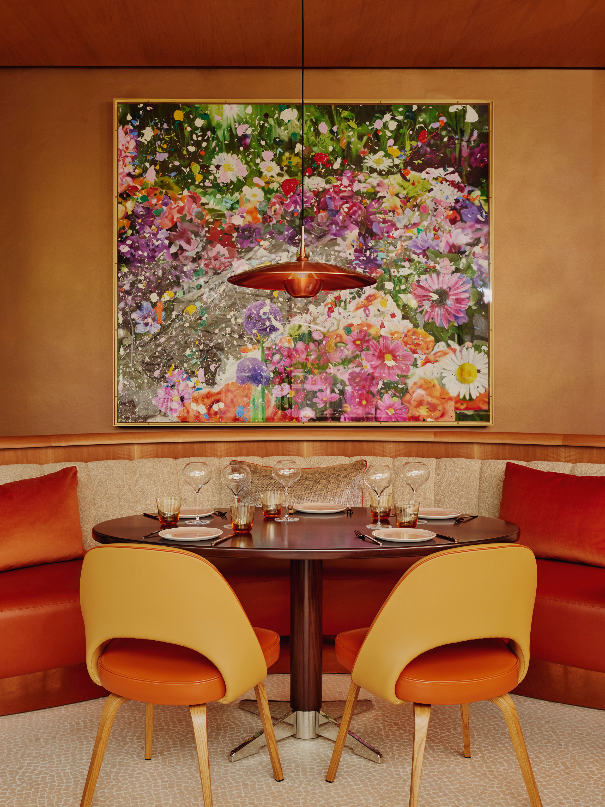 Vibrant dining nook with orange seating, glassware, and a large floral artwork.