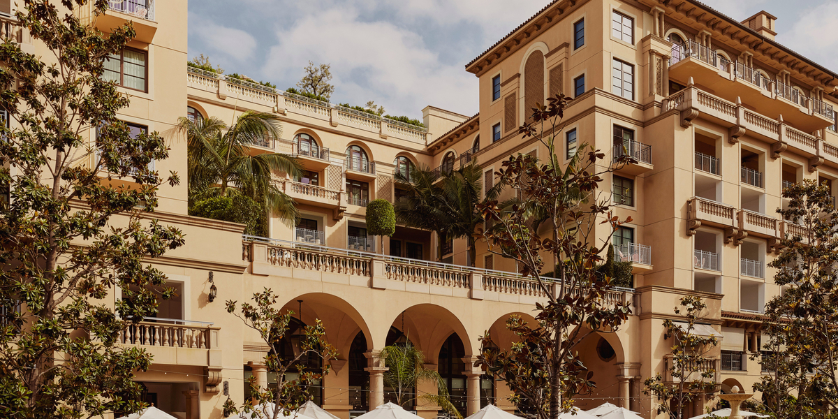 A luxurious Mediterranean-style hotel with arched balconies, lush greenery, and an outdoor terrace adorned with umbrellas and palm trees.