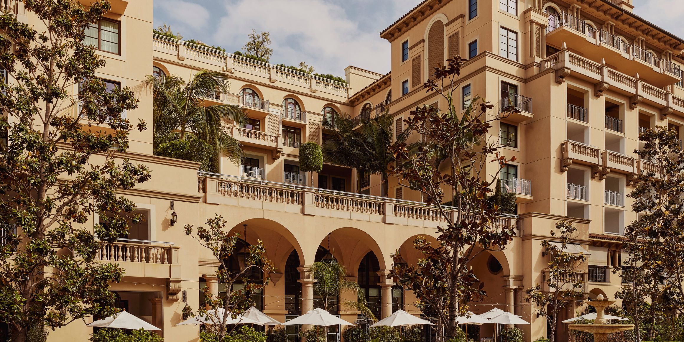Exterior of Mediterranean-style luxury hotel with arched terraces, balconies, palm trees, and white parasols in the courtyard.