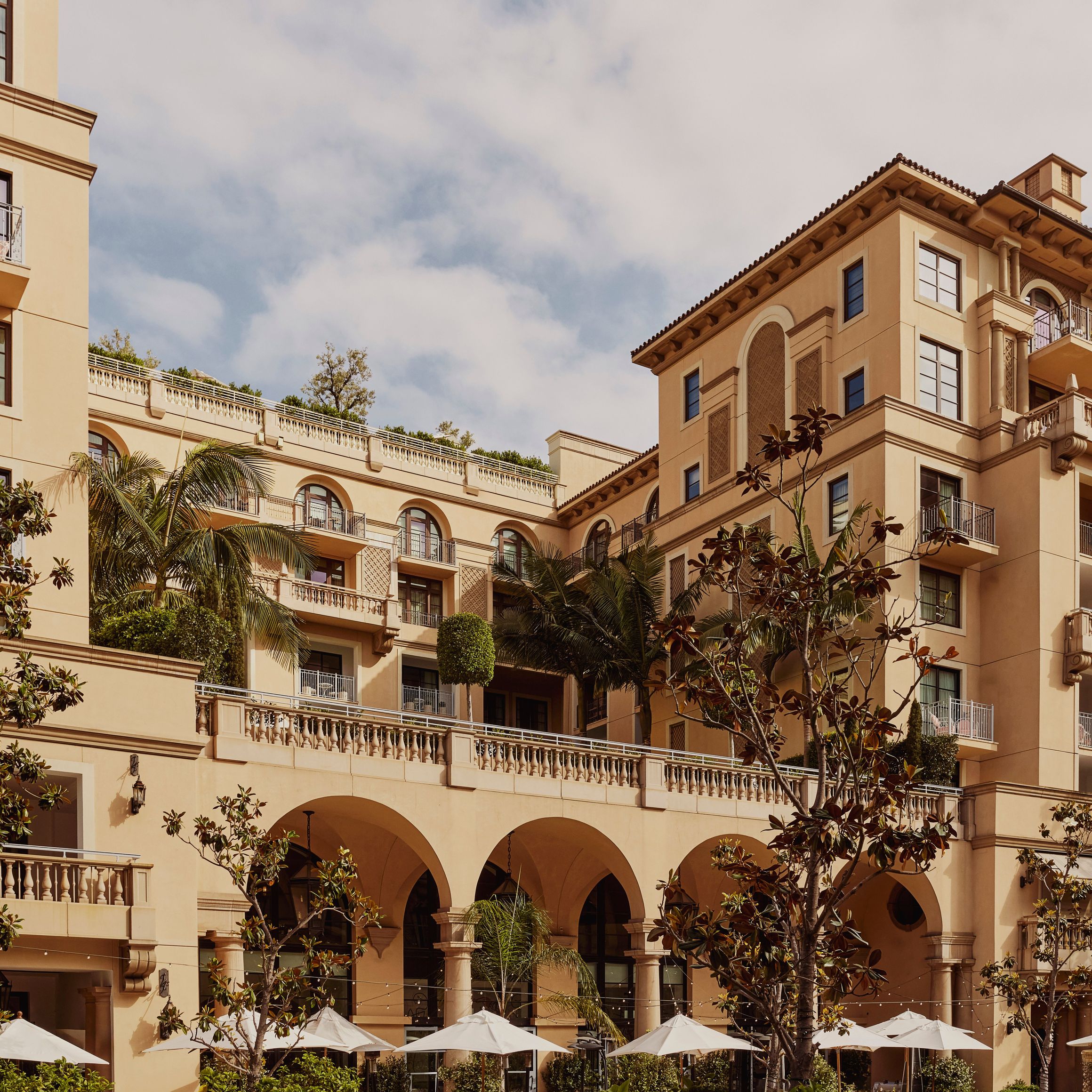 Exterior of Mediterranean-style luxury hotel with arched terraces, balconies, palm trees, and white parasols in the courtyard.
