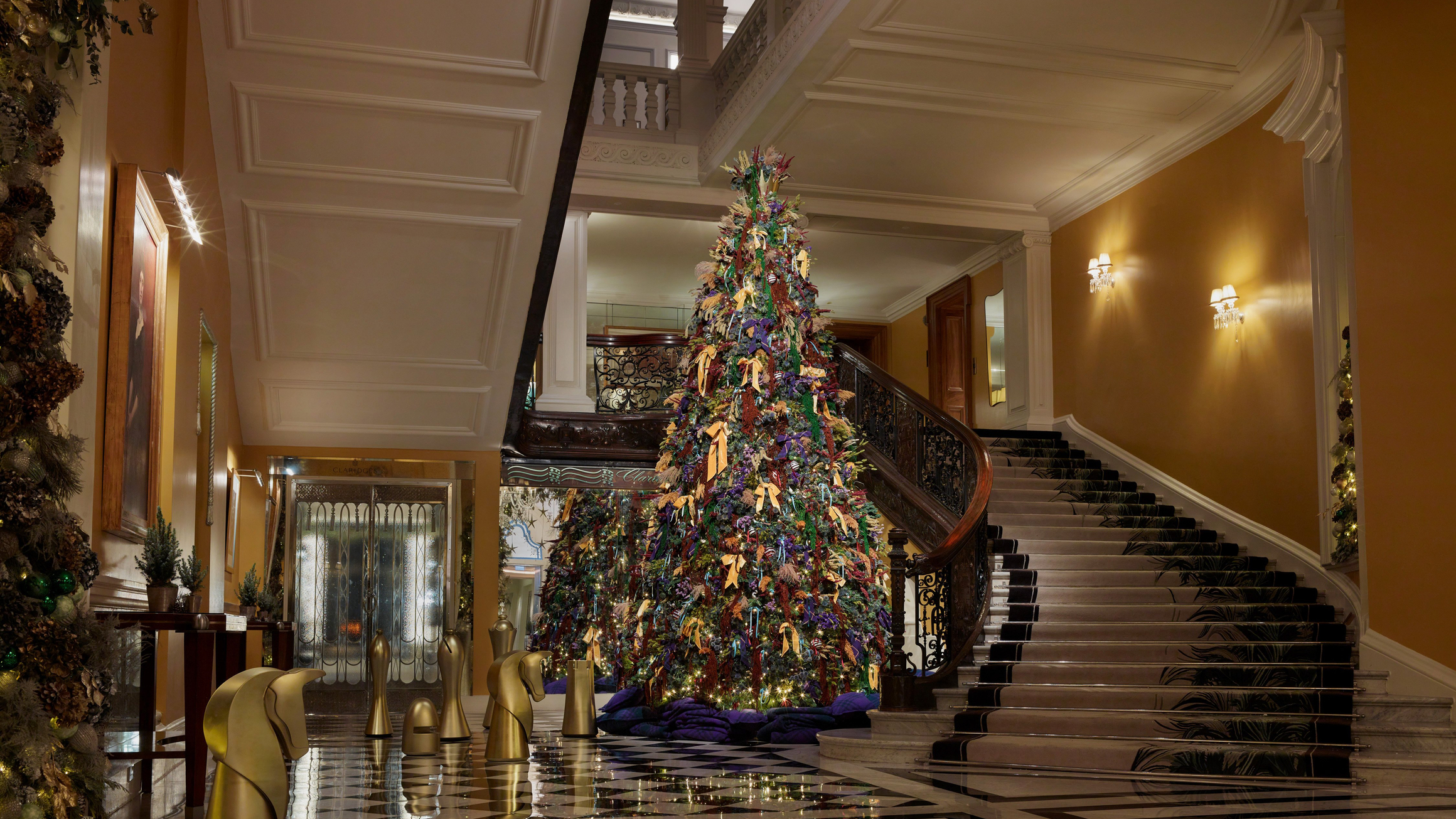Grand hotel lobby with tall decorated Christmas tree beside sweeping staircase and gold chess-piece sculptures on a marble floor.