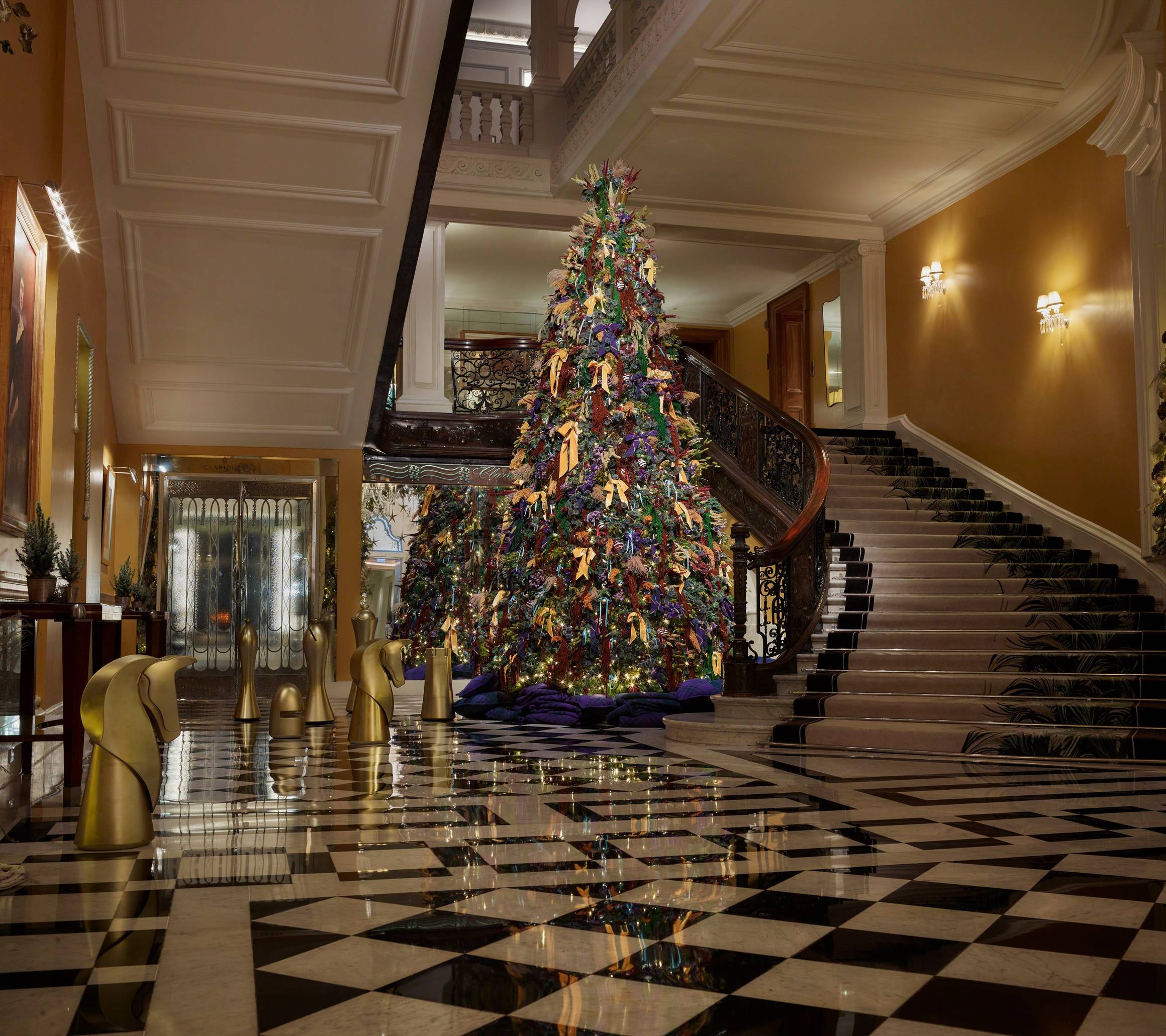 Grand hotel lobby with tall decorated Christmas tree beside sweeping staircase and gold chess-piece sculptures on a marble floor.