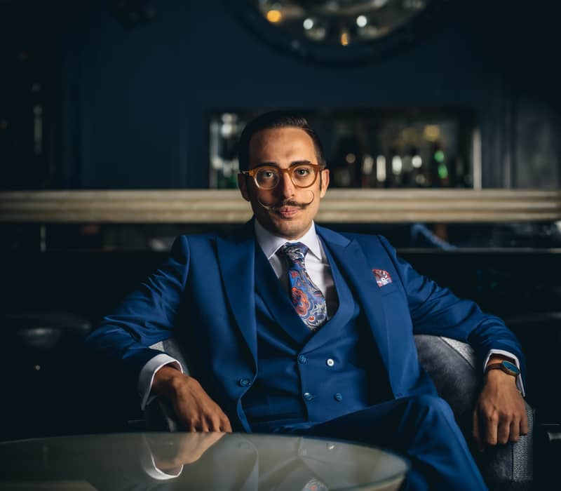 Man in a blue suit seated confidently at a cocktail table in a moody, elegant bar setting.