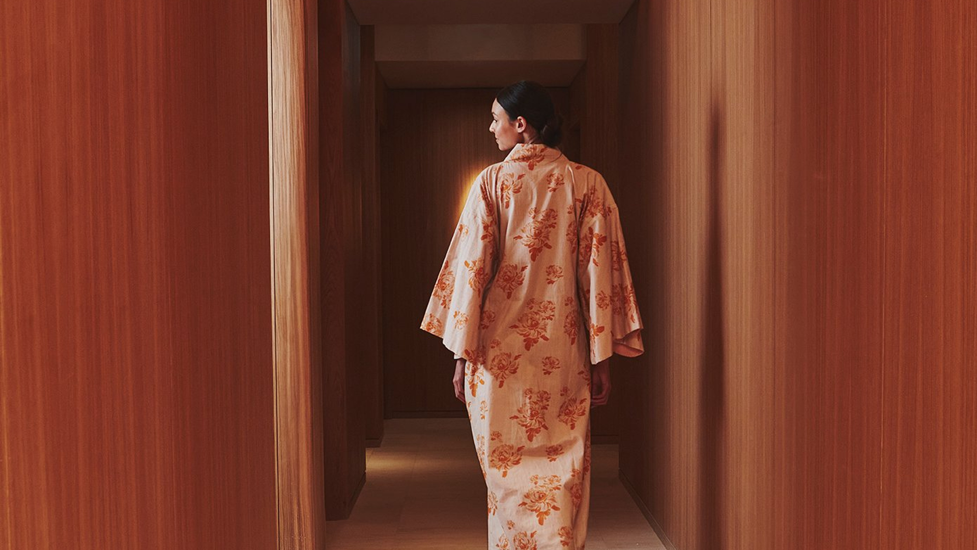 A minimalist wooden corridor with warm lighting, featuring a woman walking away gracefully in a floral kimono.