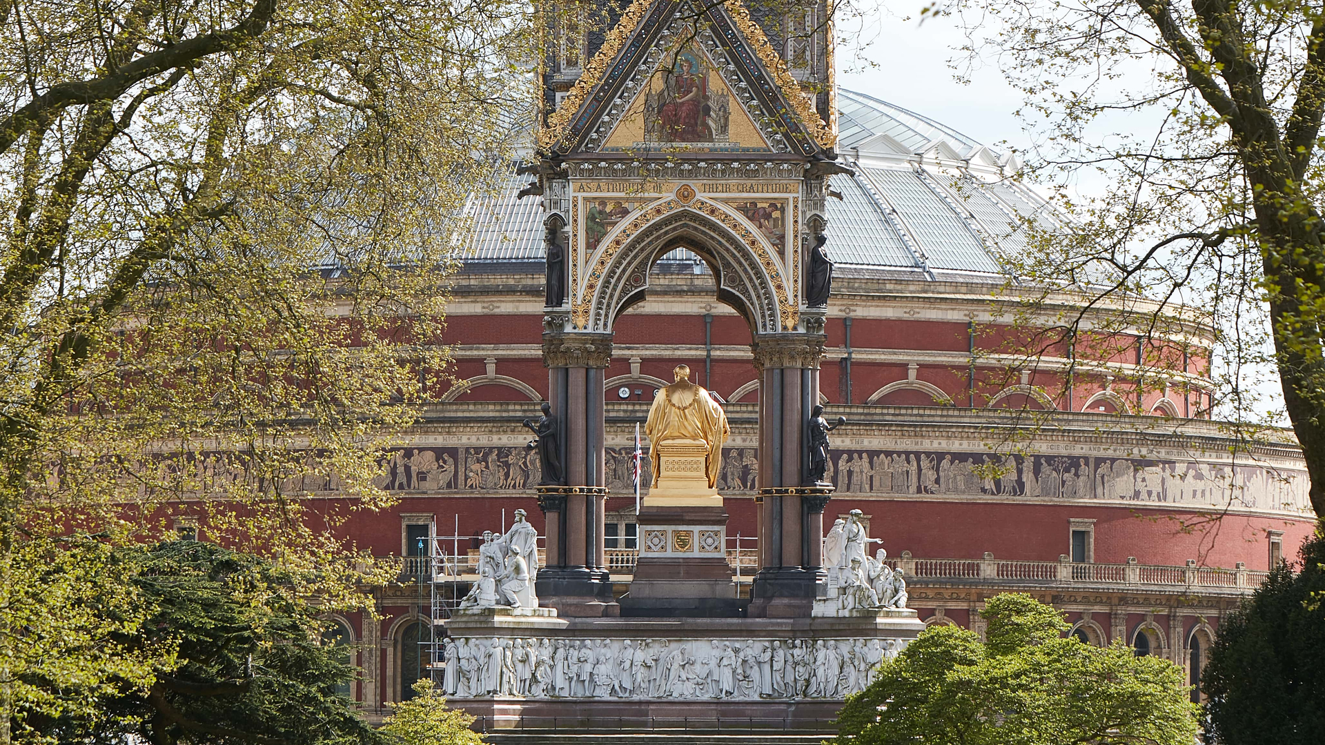 View of the Albert Memorial framed by spring foliage, with the ornate dome of the Royal Albert Hall rising behind; pedestrians and cyclists enjoy the tree-lined path in the foreground.