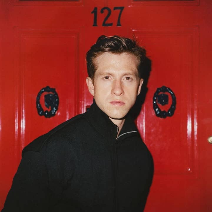 Man in a dark jacket standing in front of a bright red door marked with the number 127.