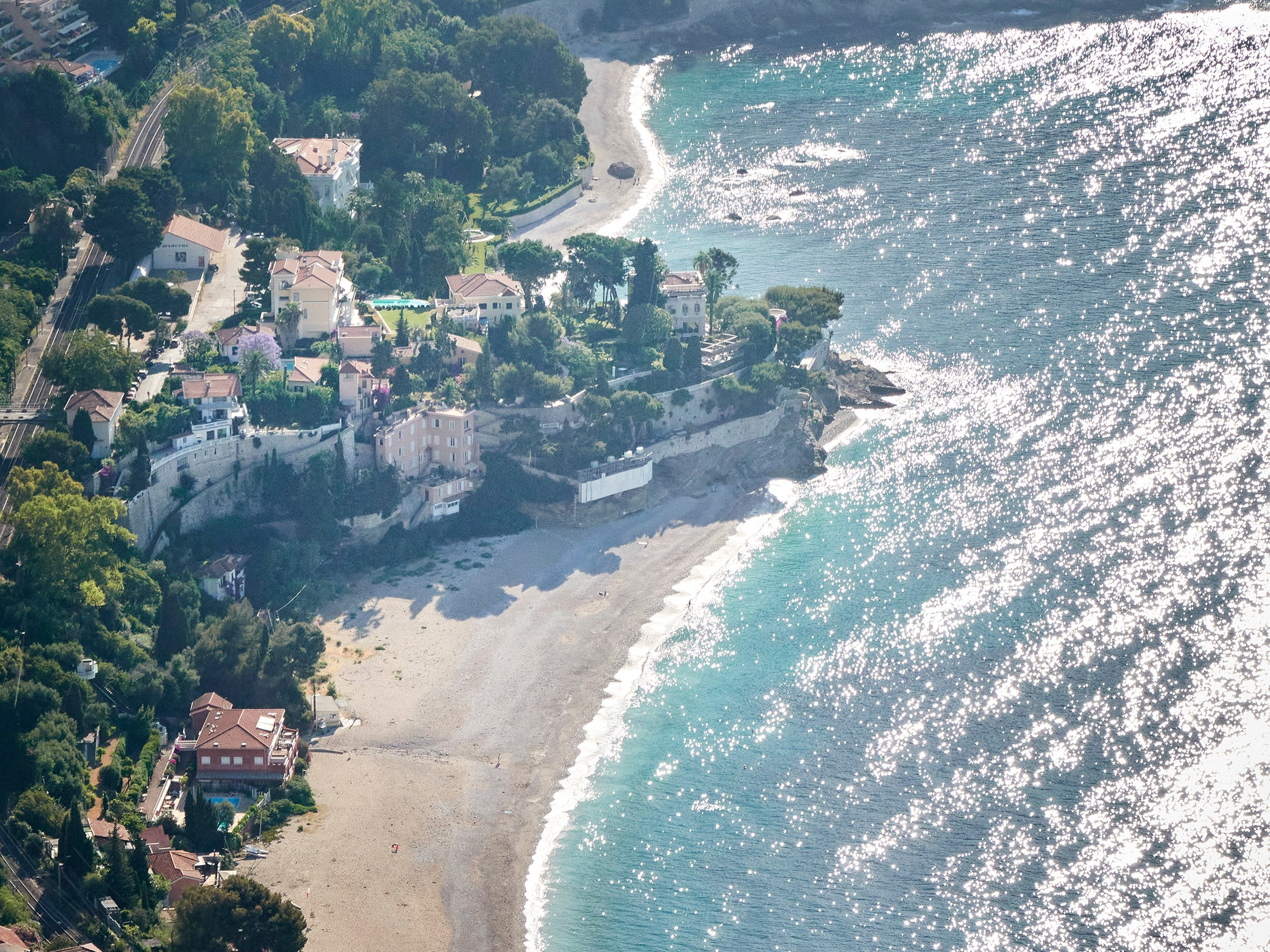 A coastal aerial view of a sunlit beach, sparkling turquoise waters, and charming villas surrounded by lush greenery.