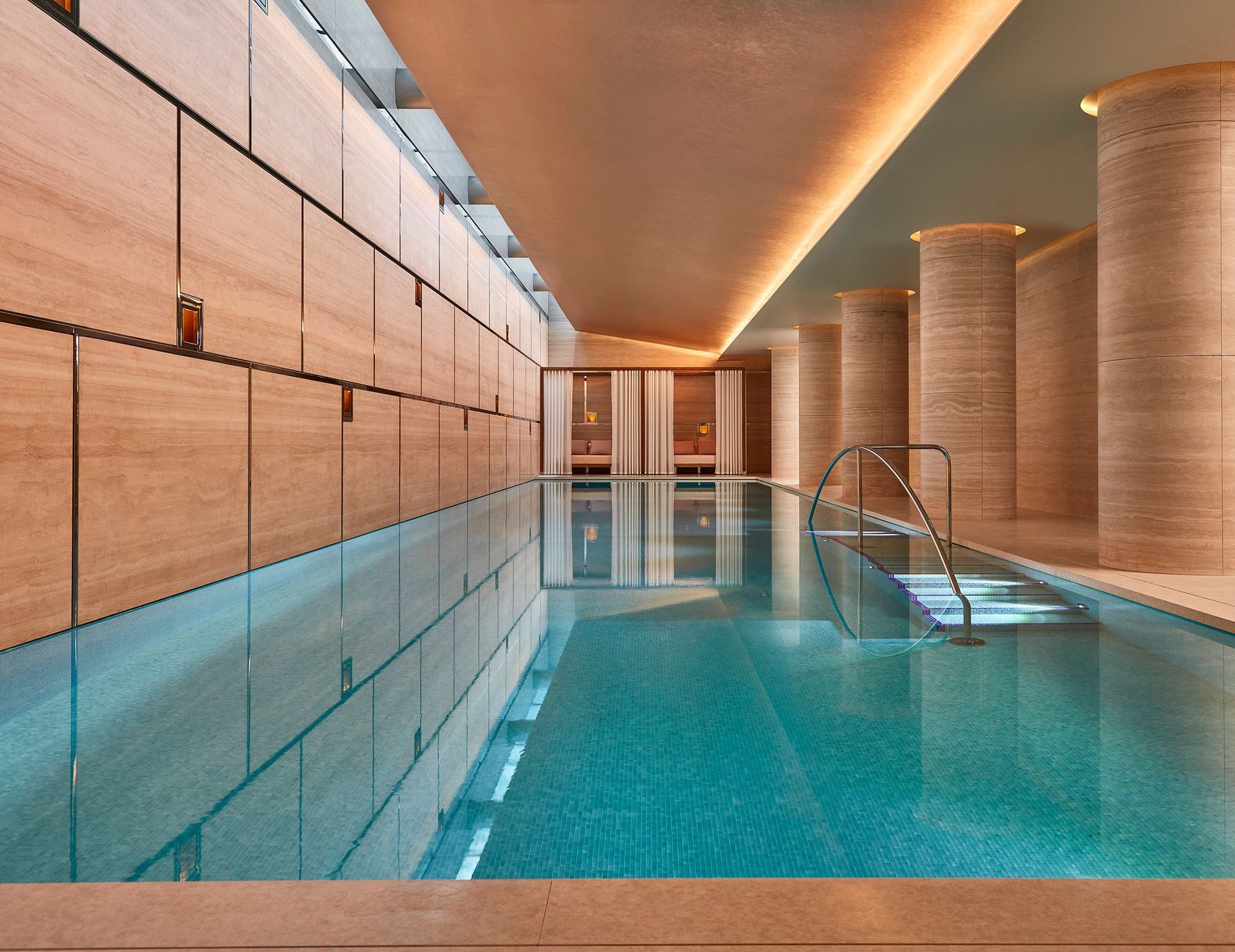 Serene indoor swimming pool with warm lighting, stone walls, and private curtained seating alcoves at the far end.
