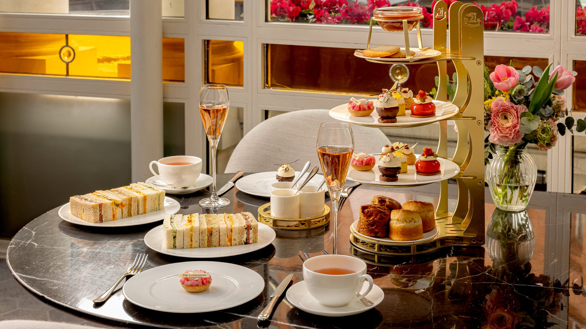 Afternoon tea set on a marble table with finger sandwiches, pastries, scones, tea, rosé champagne, and a vase of fresh flowers.