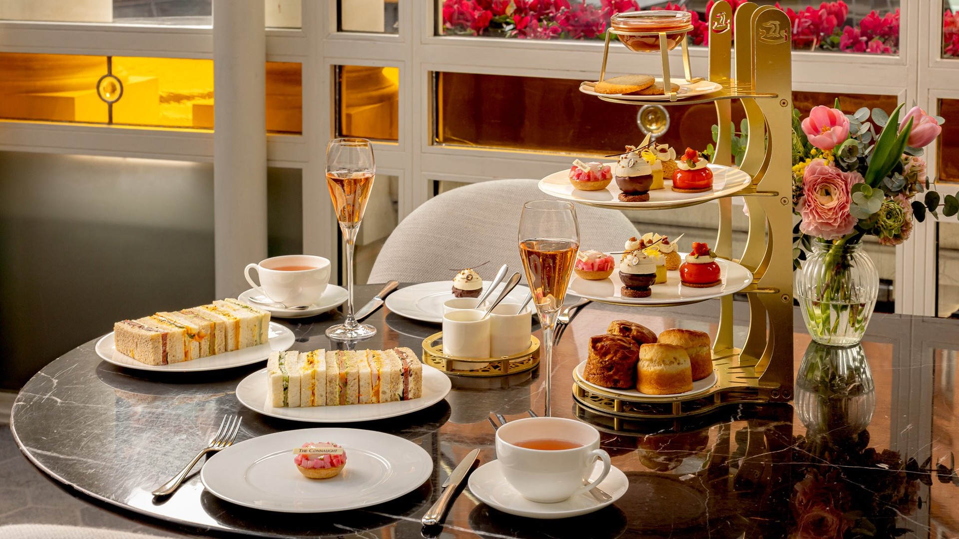 Afternoon tea set on a marble table with finger sandwiches, pastries, scones, tea, rosé champagne, and a vase of fresh flowers.