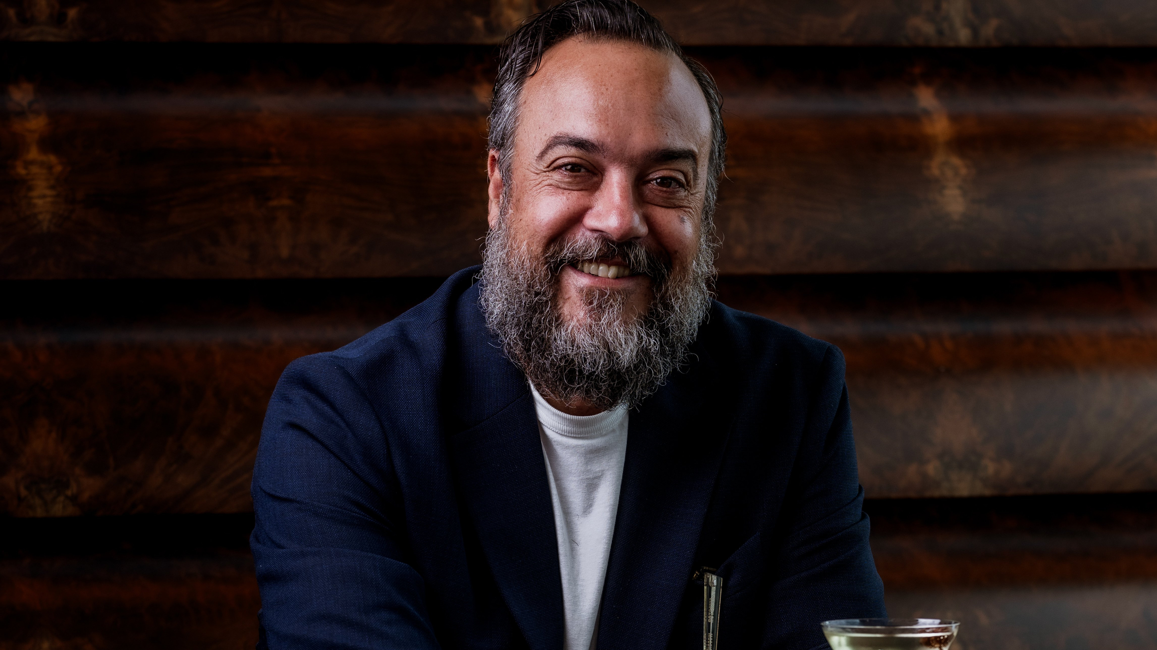A confident man in a navy suit, accessorized with bracelets, enjoys a cocktail against a warm, sophisticated backdrop.