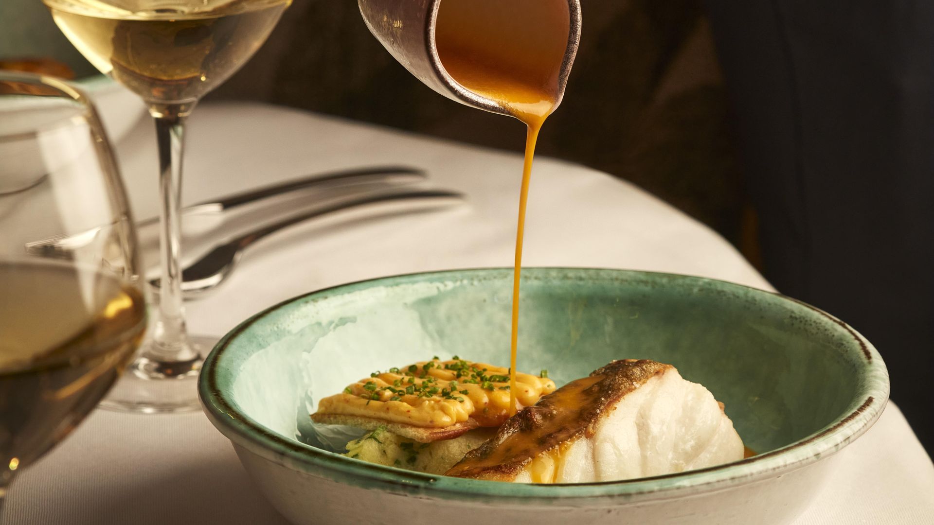 Close-up of server pouring orange sauce over seared fish and vegetables in ceramic bowl, beside two glasses of white wine.