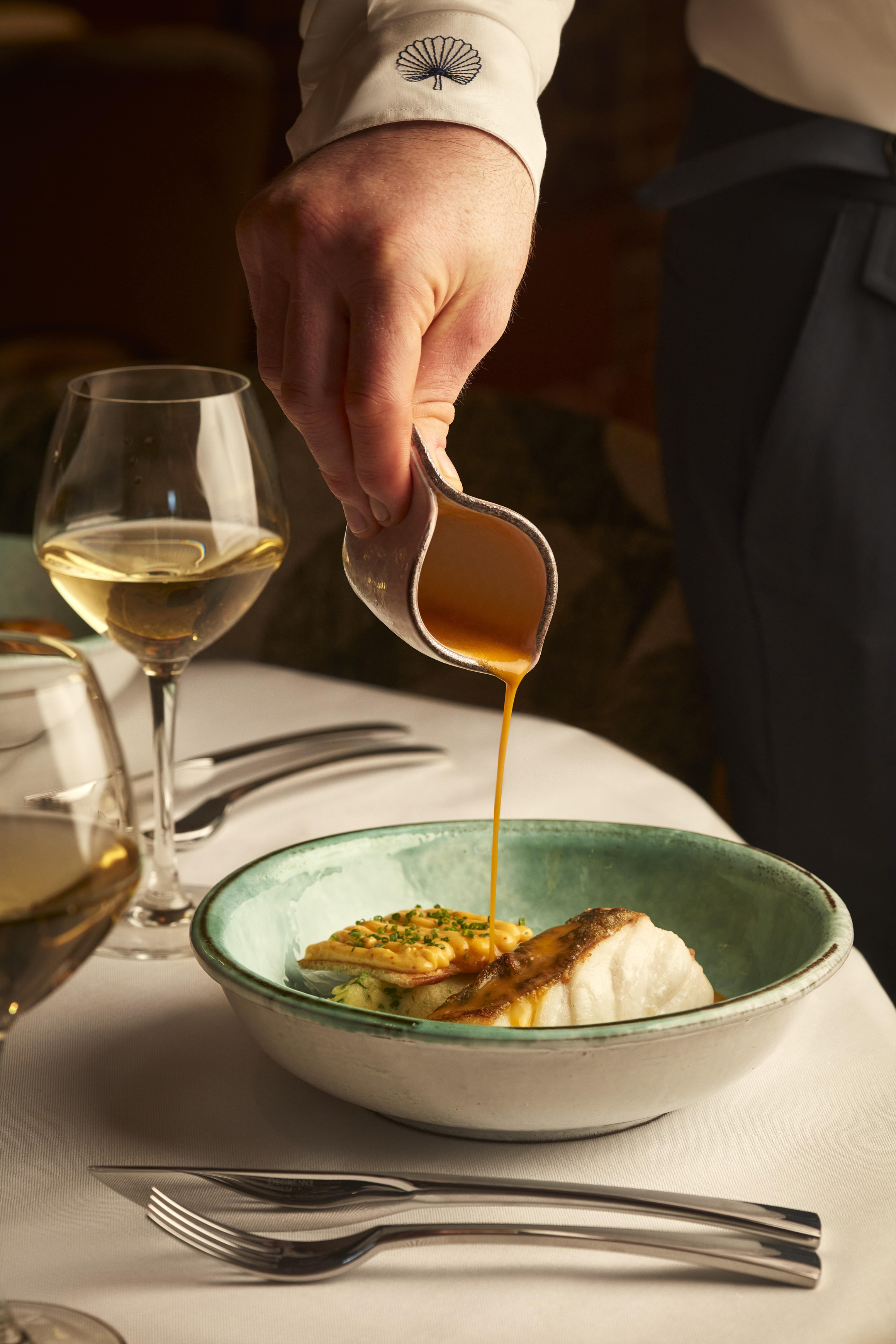 Close-up of server pouring orange sauce over seared fish and vegetables in ceramic bowl, beside two glasses of white wine.