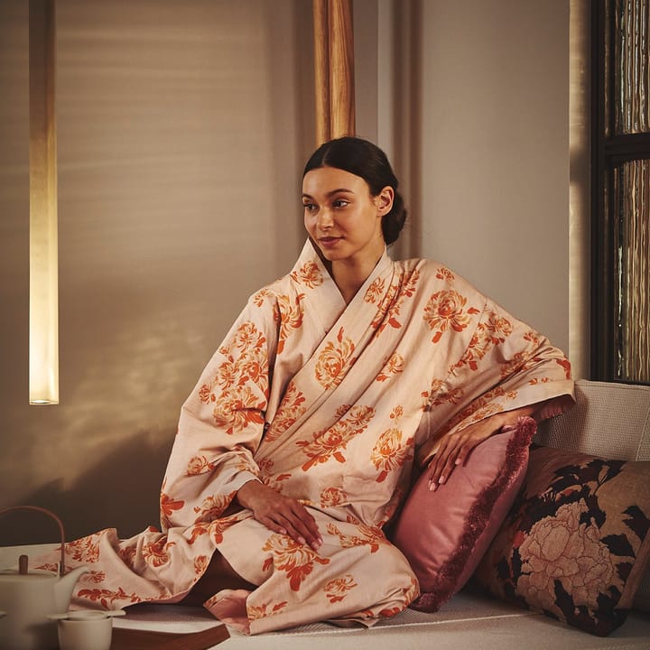 Woman in floral kimono seated on a cushioned bench in a serene spa setting, beside a tea set, surrounded by warm, ambient lighting.