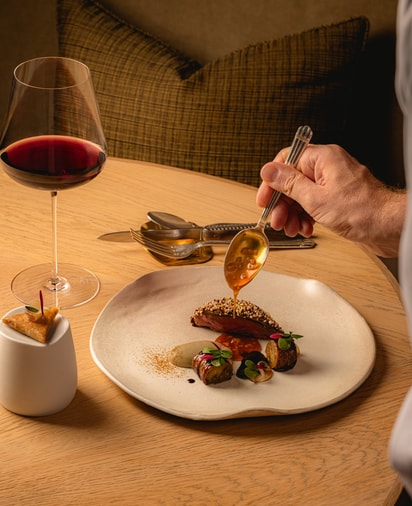 A Chef hand is carefully plating a gourmet meat dish, pouring sauce over vibrant ingredients on a round white dish, sitting on a round wooden table. A glass of red wine and cutlery are also on the table. A brown cushion and chair sit in the background.
